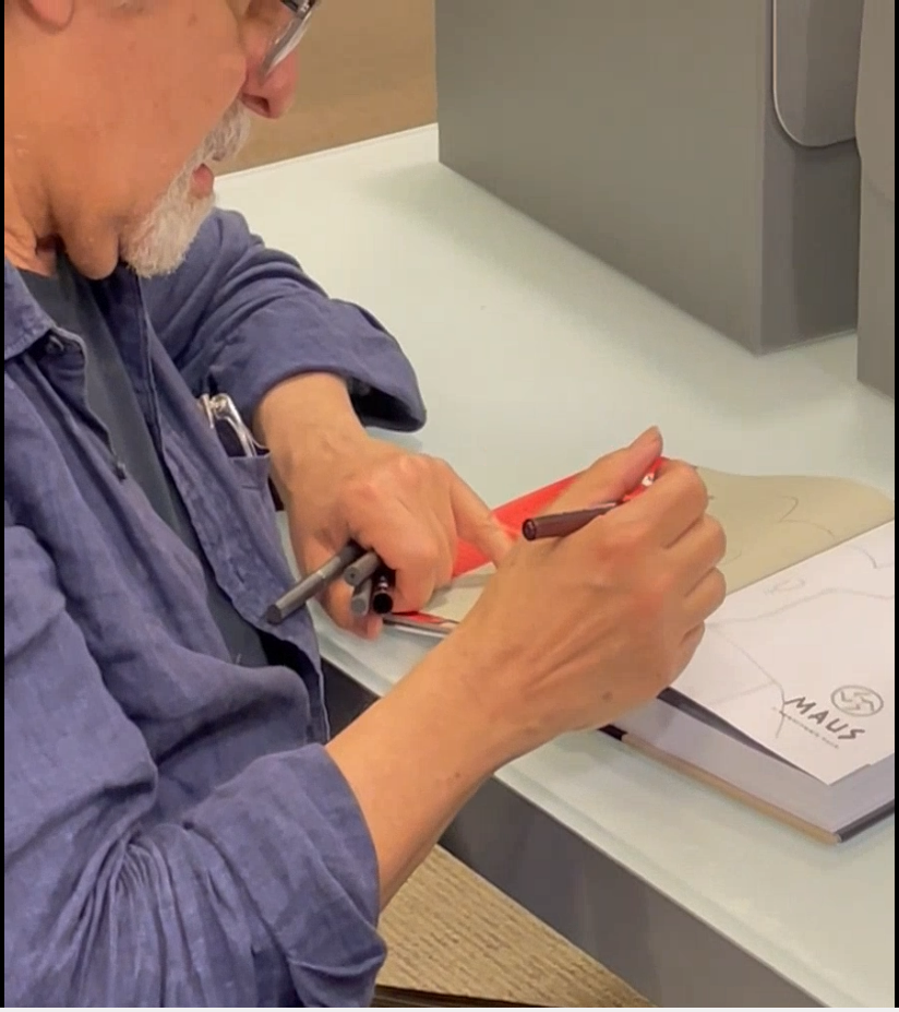 Image is of Art Spiegelman holding a pen creating an original drawing in a copy of his book Maus for Fondren Library